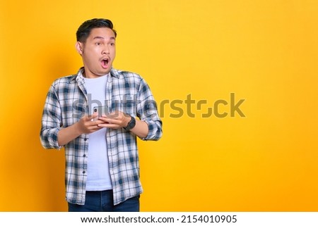 Shocked young Asian man in plaid shirt holding mobile phone and looking aside at copy space isolated on yellow background