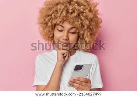 Lovely beautiful woman with curly hair watches movie online via smartphone writes message keeps hand under chin focused at cellular screen dressed in casual clothes isolated over pink background