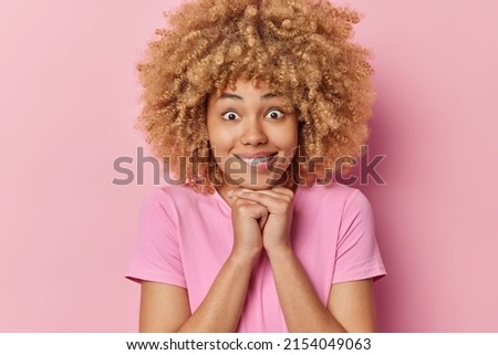 Portrait of surprised young woman with curly bushy hair keeps hands under chin bites lips has wondered expression wears casual t shirt poses against pink background. Human facial expressions Royalty-Free Stock Photo #2154049063