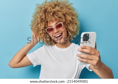 Carefree optimistic young woman has fair curly hair has cheerful expression takes selfie via smartphone wears sunglasses and casual white t shirt isolated over blue background makes video call