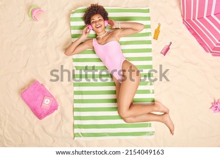 People leisure and vacation concept. Cheerful curly haired beautiful woman dressed in swimwear enjoys favorite playlist while lying in sun at beach has lazy day unforgettable summer holidays
