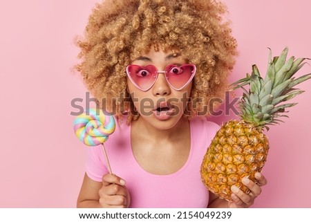 Beautiful shocked woman with curly hair wears heart pink sunglasses holds lollipop and pineapple chooses between healthy and unhealthy food stares anxious at camera isolated over pink background