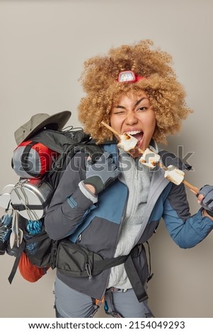 Vertical shot of female tourist eats tasty marshmallow roasted on bonfire has picnic and adventure carries rucksack dressed in comfortable clothes isolated over grey background. Pastime recreation