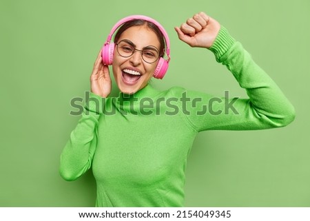 Positive good looking young woman with dark hair moves carefree catches every bit of song uses stereo headphones wears round spectacles casual turtleneck isolated over vivid green background