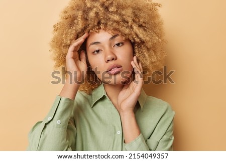 Portrait of serious woman with curly hair touches face gently has healthy clean skin looks directly at camera tenderly wears green shirt poses against brown background. Natural beauty concept