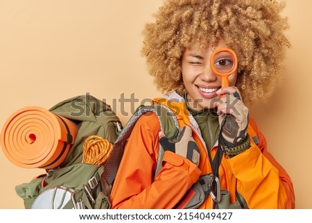 Positive curly female tourist smiles happily holds magnifying glass over eye explores new place carries backpack with necessary items for travel isolated over brown background. Woman backpacker Royalty-Free Stock Photo #2154049427