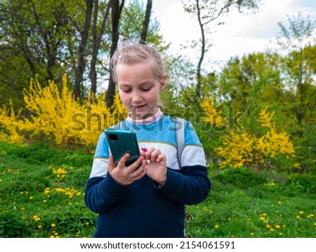 Adorable child girl chatting talking in social media smartphone learning language reading books outdoor. Blogger kid with gadget distance school online podcast lesson cell phone in park yellow flowers