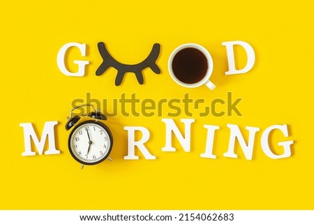 Creative good morning concept. White letters, black eyelashes, alarm clock and cup of coffee on yellow background. Top view, Flat lay. Greeting card.