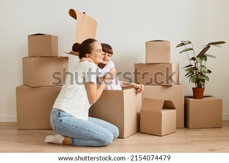 Image of little girl with her mother sitting in handmade boat from cardboard boxes and hugging, happy child playing with mommy during moving in a new flat.