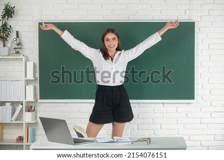 Portrait of a young, confident and attractive female student excited amazet student with raised hands in school classroom on blackboard background.