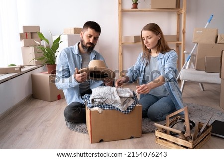 a young couple in a new apartment on day of the move sit on floor unpack things a man holds a hat in his hands. The couple moves into a new apartment. Lots of packing boxes around them. copy space.