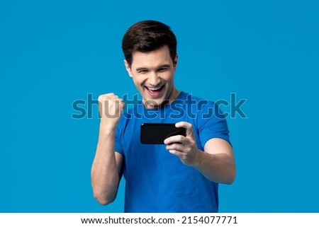 Handsome young male gamer clenching his fist with smiling face while playing game on mobile phone in isolated blue studio background