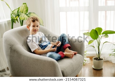 A cheerful boy with a phone in his hands is sitting in a chair and playing a game or watching cartoons.