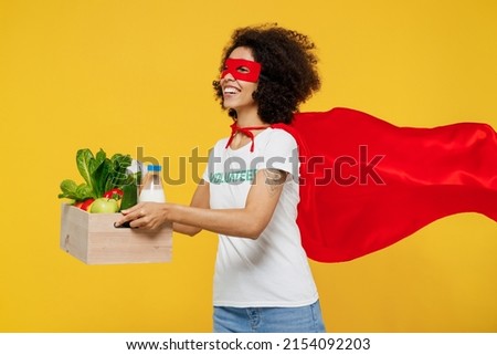 Young side view woman of African American ethnicity in white volunteer t-shirt super hero costume hold food box isolated on plain yellow background. Voluntary free work assistance help grace concept