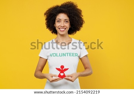 Young woman of African American ethnicity wears white volunteer t-shirt hold in hands little paper angel isolated on plain yellow background. Voluntary free work assistance help charity grace concept