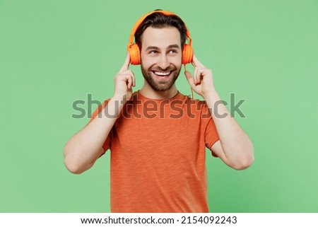 Young smiling fun satisfied cool man 20s wear casual orange t-shirt headphones listen to music dancing isolated on plain pastel light green color background studio portrait. People lifestyle concept