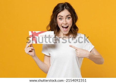 Young surprised shocked fun excited happy caucasian student woman 20s wearing white basic t-shirt point index finger on gift voucher flyer mock up isolated on yellow orange background studio portrait