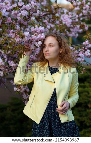 A redhead woman in a yellow jacket is looking aside, standing near a blooming magnolia. The calm woman is enjoying nature.