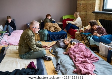 Mature man with open book reading fairy tale to his grandson lying in bed while resting in temporary shelter with other refugees Royalty-Free Stock Photo #2154115639