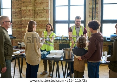 Several refugees standing by table with cooked food in front of two volunteers putting dinner into containers and passing them to migrants Royalty-Free Stock Photo #2154115725