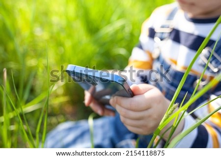 The boy sits in the backyard in the grass and watches cartoons on his phone. People use technology, summer holiday