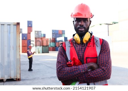 Portrait of African American young engineer worker man wearing safety bright neon red color vest and helmet, standing with arms crossed at logistic shipping cargo container yard as blurred background.