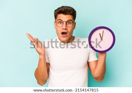 Young caucasian man holding a clock isolated on blue background surprised and shocked.