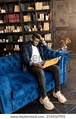 Serious African american manager wear headphones looking at laptop screen provide support to client distantly, hold online conference. Student study distantly at modern blue and yellow apartment.