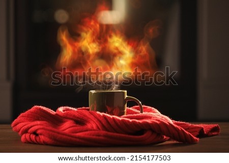 Cup of hot drink and red knitted scarf on wooden table near fireplace at home. Cozy atmosphere Royalty-Free Stock Photo #2154177503