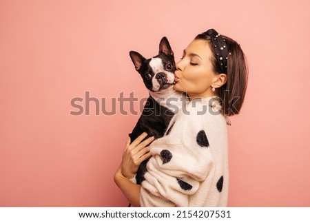Light-skinned young brunette woman kisses her pet Boston Terrier hard on pink background. Girl in white sweater loves dogs very much. True friendship, wonderful happy moments