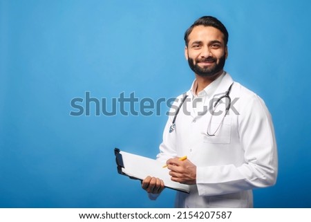 Indian doctor in white medical gown with stethoscope on shoulders taking notes standing with clipboard isolated on blue, physician therapist writing down treatment plan, writes a prescription