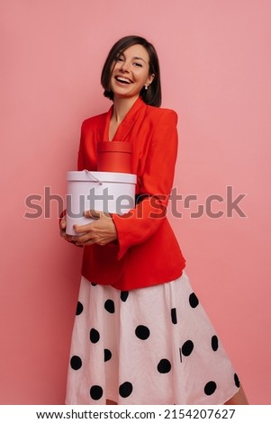 Cute young caucasian girl smiles, holds gift box in her hands on pink background. Brunette with short haircut wore red jacket and white skirt for occasion. Celebration concept