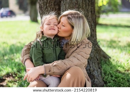 woman kissing happy daughter while sitting under trees in park