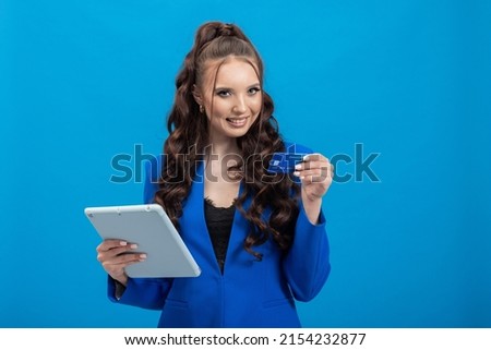 Photo of fashionable brunette holding plastic credit card and tablet, online payment in store, ordering clothes, entering security code, isolated on blue background.