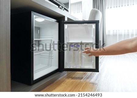 Man Hand opened mini small refrigerator and reached drinking plastic water bottle under the wood counter in the hotel resort bedroom. Royalty-Free Stock Photo #2154246493