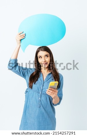 Dreamy brunette girl using smartphone and holding empty speech bubble looking up while standing on white background