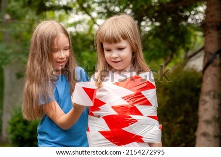 Girl wrapping her hyperactive sister in red white safety tape, sisters, siblings, friends playing. Children safety, kids protection, ADHD, help, overprotectiveness, restraint, protection concept Royalty-Free Stock Photo #2154272995