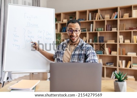 E-learning concept. Excited male English teacher sitting at table, pointing with pen at whiteboard, explaining grammar rules to students, having online lesson from home Royalty-Free Stock Photo #2154279569