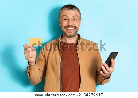 Mobile Shopping Application. Middle Aged Man Holding Cellphone Showing Credit Card Smiling To Camera Standing Over Blue Background. Studio Shot Of Happy Buyer. Ecommerce Concept