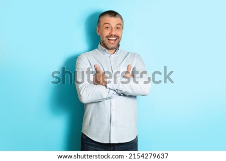 Cheerful Mature Man Laughing Smiling Looking At Camera Standing Crossing Hands Over Blue Studio Background. Male Wearing Shirt Posing Expressing Positive Emotions