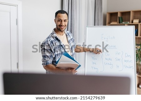 Online tutoring. Happy male teacher giving English class, pointing at blackboard with basic grammar rules and smiling at laptop webcam. Experienced professor explaining new material to students Royalty-Free Stock Photo #2154279639