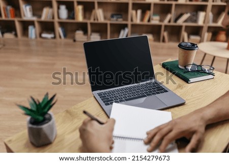 Online class concept. Male student writing in notebook, taking notes and using laptop with black screen for mockup template, watching lecture or webinar at home