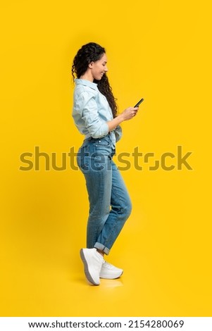 People And Technology Concept. Portrait of happy smiling young woman using smartphone standing isolated on yellow orange wall. Female chatting online, full body length, profile side view portrait