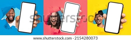 Wow, Great Offer. Three excited young diverse people holding 3 big cell phones with white blank screen in hand, showing to camera through torn paper holes. Gadgets with empty free copy space, banner Royalty-Free Stock Photo #2154280073