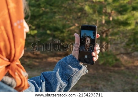 An elderly woman takes photo of selfie on mobile phone while walking in a pine forest. Grandma communicates via video communication in a smartphone. Modern old lady in a denim jacket and a headscarf.