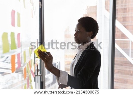 Focused confident Black business leader woman preparing project plan presentation, sticking colorful paper notes on glass board, working on tasks, strategy, using scrum management tool. Royalty-Free Stock Photo #2154300875