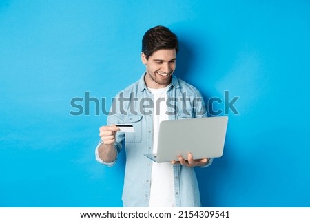 Young smiling man buying in internet, holding credit card and paying for purchase with laptop, standing over blue background
