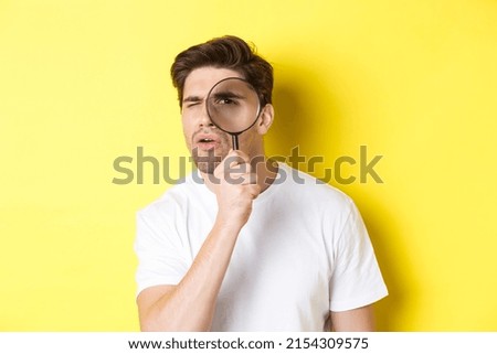 Man looking through magnifying glass with serious and thoughtful look, searching or investigating, standing over yellow background