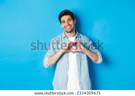 Handsome man smiling, showing heart gesture and looking at camera, saying I love you, standing against blue background