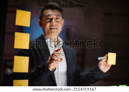 Close up blurred view of smiling handsome Asian businessman, standing behind of office glass wall, using post it notes while planning project tasks in modern office at night. Focus on hand